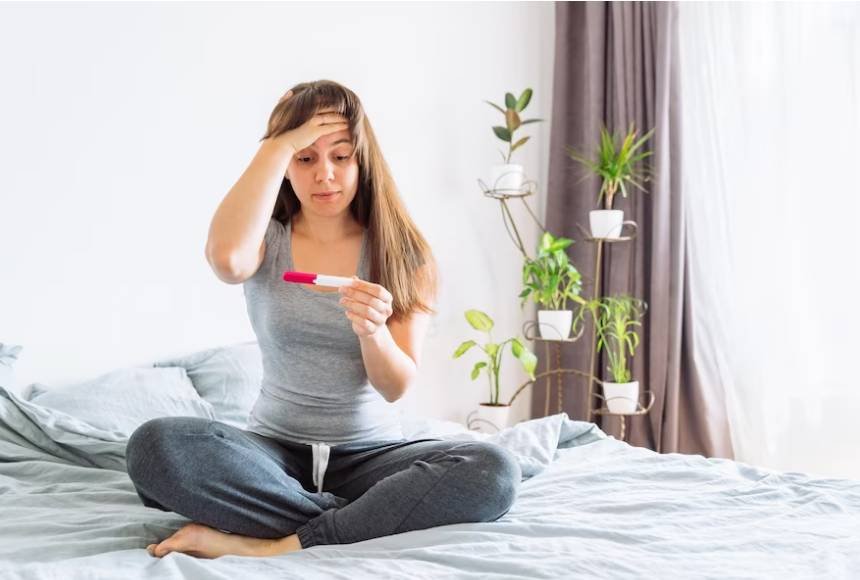 A young woman sitting on her bed, looking at a positive pregnancy test with a worried expression.