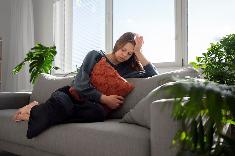 A woman sitting on a sofa holding a pillow, appearing to be in discomfort.